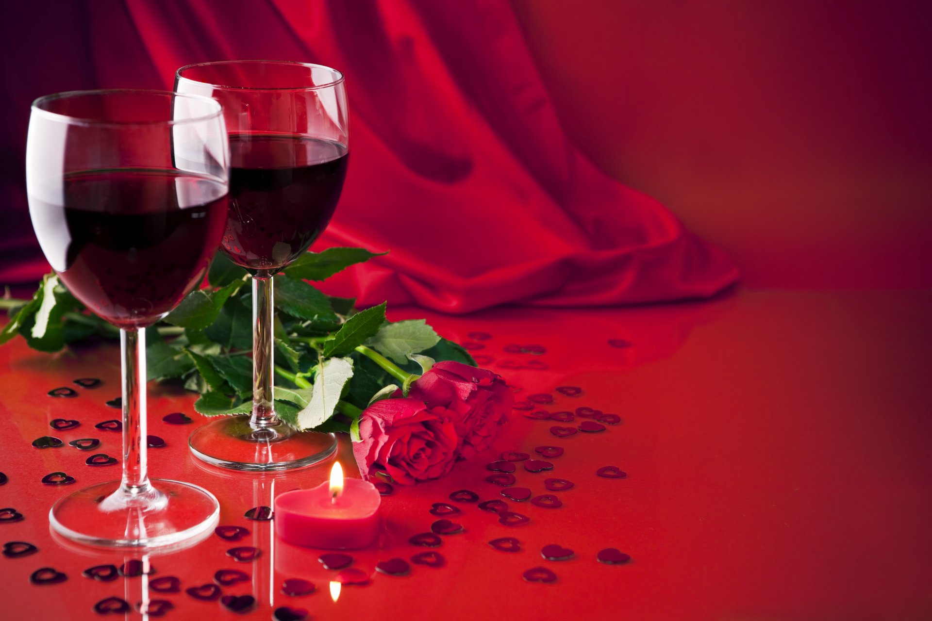 Two glasses of red wine beside a red rose, scattered heart-shaped confetti, and a lit candle on a red surface, captured in 4K Ultra HD for a PC desktop wallpaper.