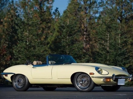 HD PC desktop wallpaper featuring a classic cream-colored Jaguar E-Type convertible parked in front of a forested background.