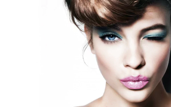 A glamorous model with blue eyes winks playfully, showcasing bold lipstick on her lips against a striking white background, reflecting a stylish Hungarian celebrity vibe.