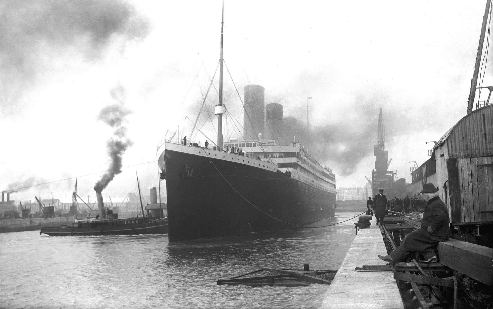 2K Quad HD PC desktop wallpaper: black-and-white image of the RMS Titanic docked, tugboats and crew along an industrial harbor with smoke pouring from the ship's funnels.