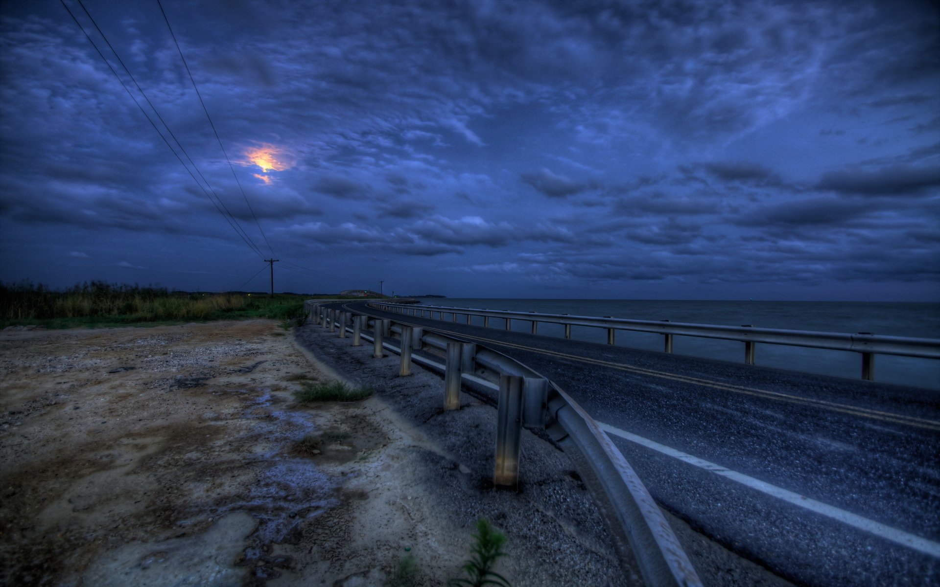 A serene HD desktop wallpaper depicting a winding road beside the ocean, under a dramatic sky filled with clouds and a glowing moon, showcasing a blend of man-made and natural elements.