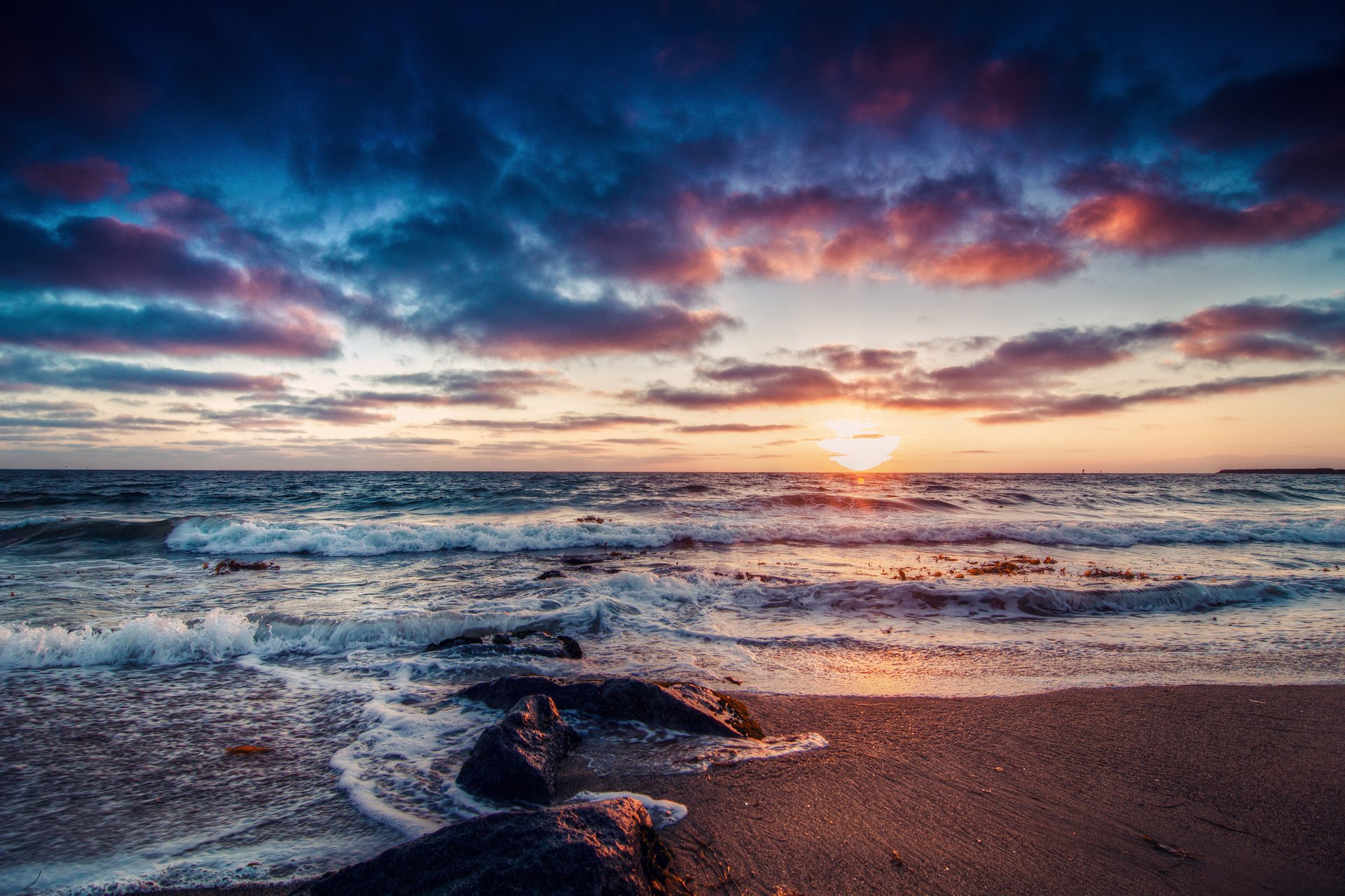 A stunning sunset over a tranquil ocean, with colorful clouds reflecting on the water. This HD nature wallpaper captures the serene beauty of the beach at dusk.
