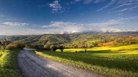 HD desktop wallpaper featuring a winding man-made road through vibrant green fields and rolling hills under a bright blue sky with scattered clouds.