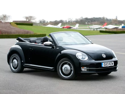 2013 black Volkswagen Beetle Cabriolet 50s Edition convertible parked on an airfield, with small planes and green fields in the background.