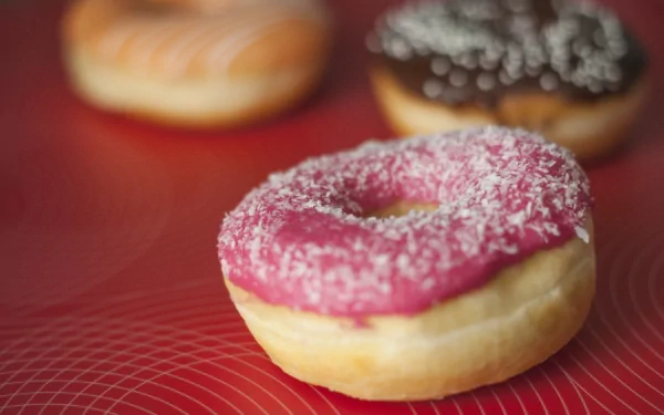 A vibrant pink doughnut topped with coconut flakes stands out against a red background, complemented by other doughnuts in various flavors. A delicious food-themed 4K Ultra HD wallpaper.