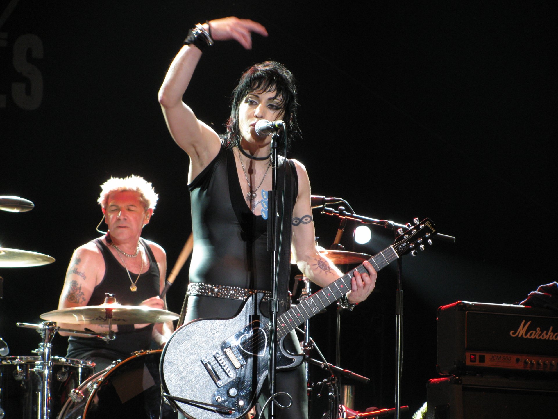 HD PC desktop wallpaper featuring Joan Jett performing live with her guitar, backed by a drummer on stage, capturing a dynamic music moment.