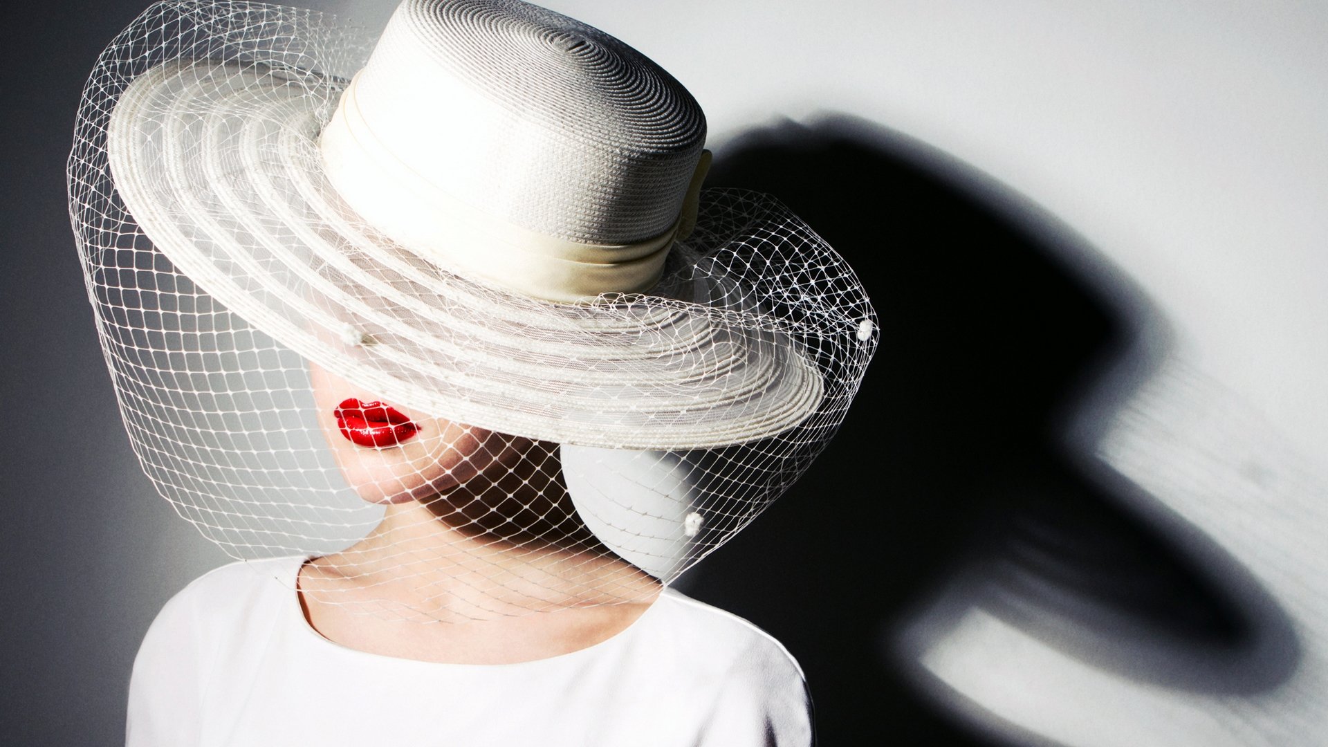 A woman model wears a wide-brimmed white hat with a veil, featuring bold red lips. The striking shadow adds depth to this HD desktop wallpaper and background.