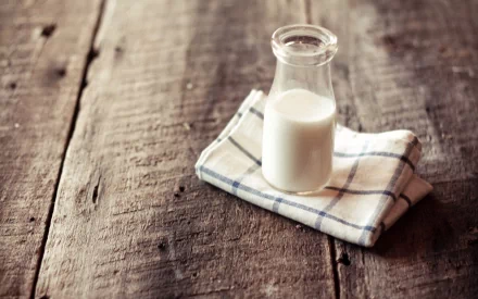 A high-definition desktop wallpaper of a small glass bottle filled with milk, placed on a folded plaid cloth on a rustic wooden table.