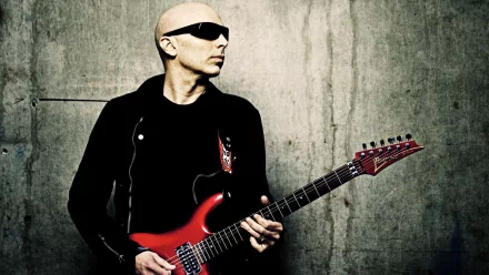 A striking HD wallpaper featuring Joe Satriani, a renowned guitarist, passionately holding his iconic red guitar against a textured concrete backdrop.