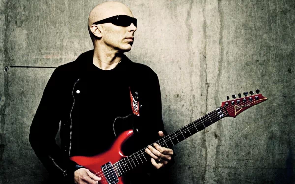 A striking HD wallpaper featuring Joe Satriani, a renowned guitarist, passionately holding his iconic red guitar against a textured concrete backdrop.