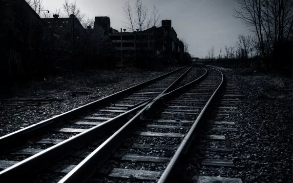 A black and white HD wallpaper featuring a railroad track curving into the distance, surrounded by sparse trees and urban structures, creating a moody and atmospheric scene.