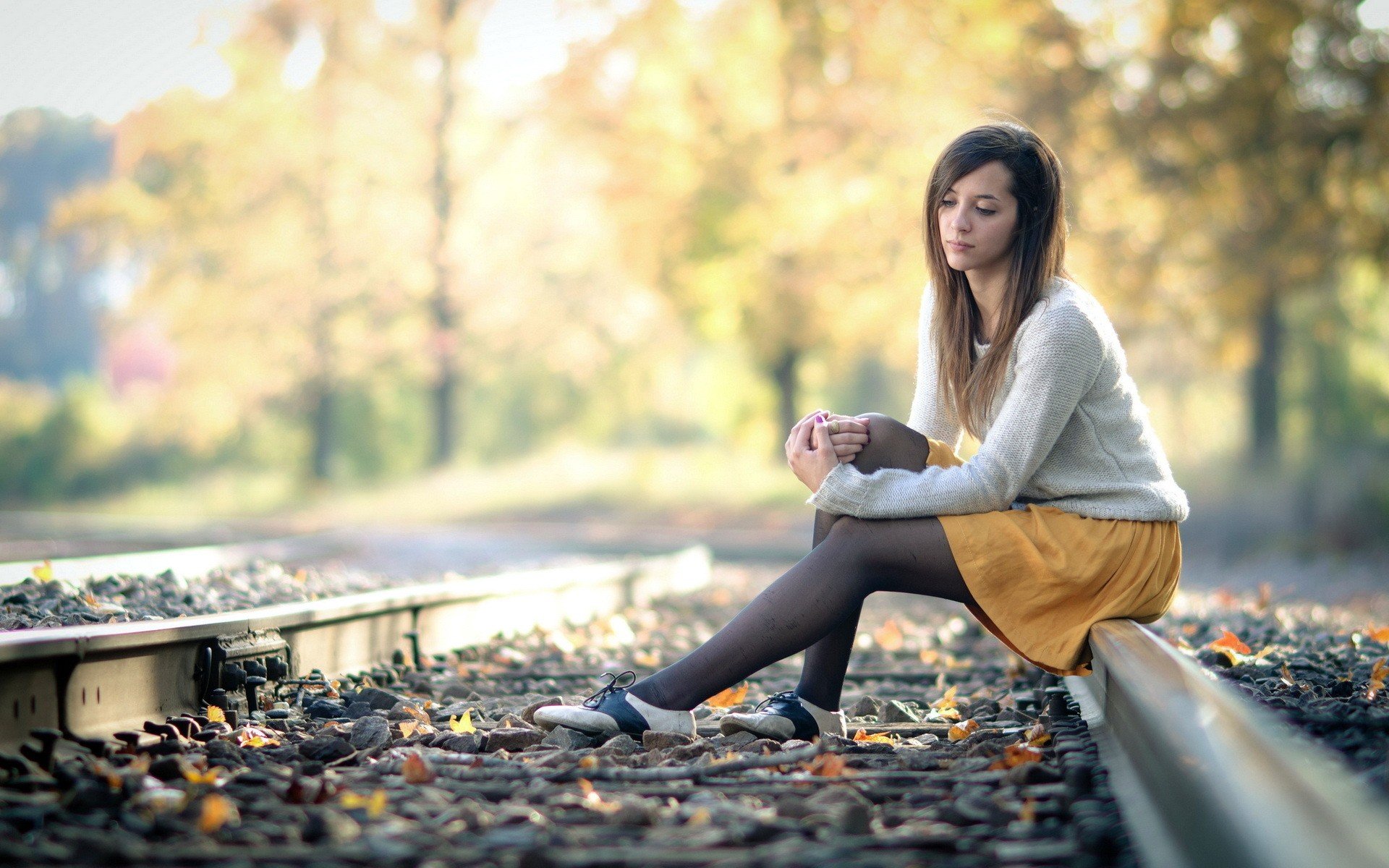 A beautiful woman sits thoughtfully on a railway track amid an autumn forest. This HD desktop wallpaper exudes a serene and reflective atmosphere with warm, earthy tones.