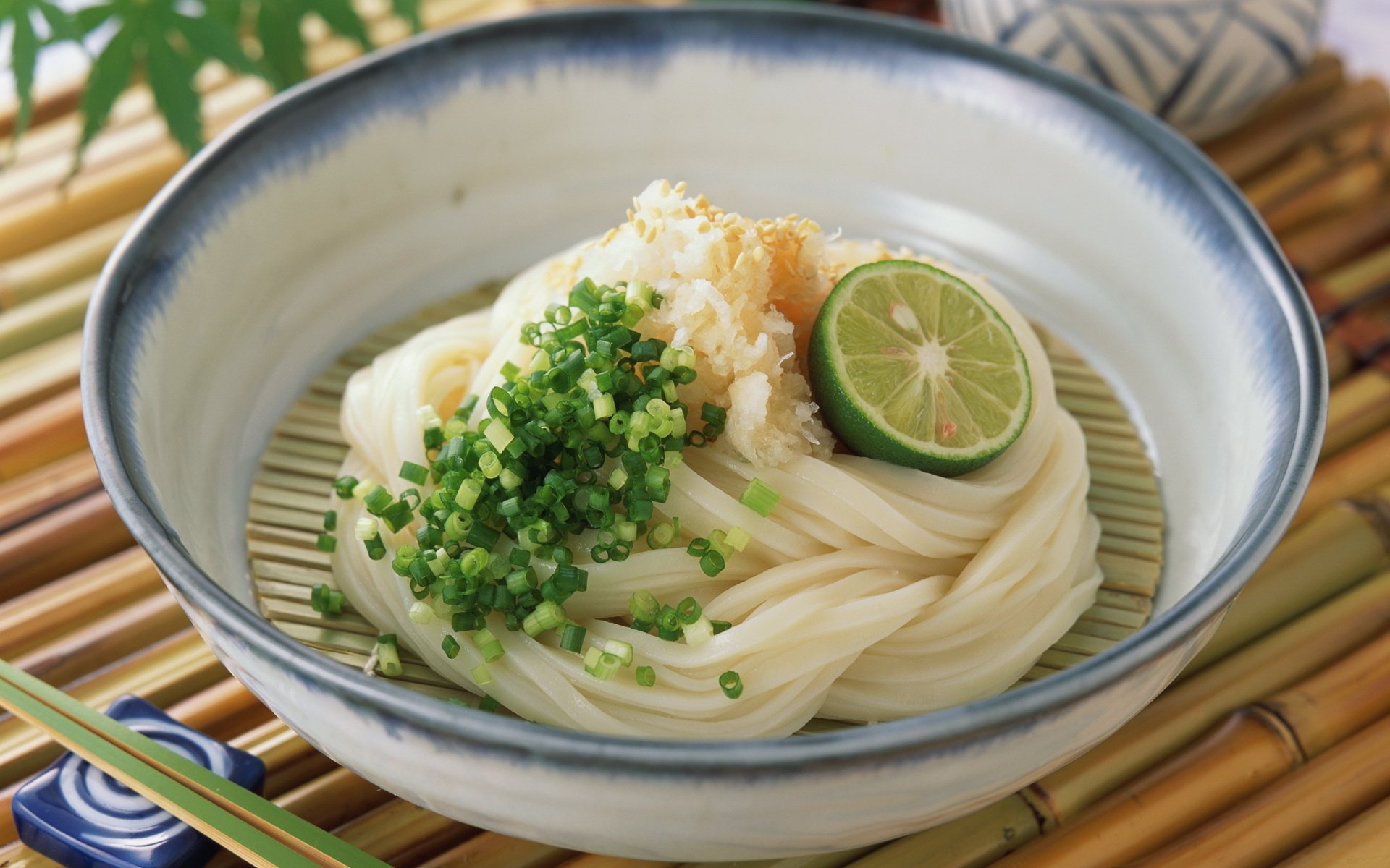HD PC desktop wallpaper of a bowl of cold udon pasta topped with chopped green onions, grated radish, and a lime slice, set on a bamboo mat with chopsticks.