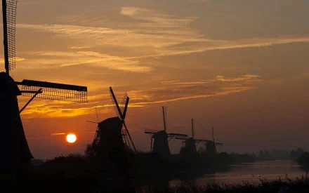 HD desktop wallpaper of man-made windmills silhouetted against a golden sunset sky over calm waters.
