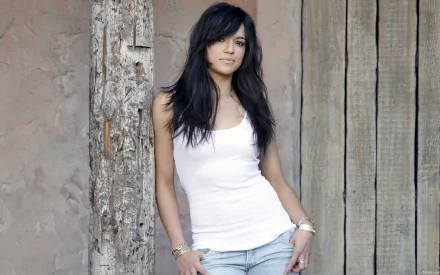 Michelle Rodriguez poses confidently against a rustic wooden backdrop, wearing a white tank top and jeans. This HD image serves as a striking desktop wallpaper.