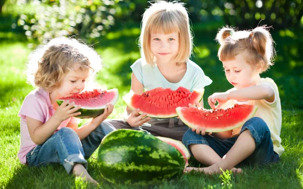 Three cheerful children sit on green grass, enjoying fresh watermelon slices in this vibrant HD photograph, making it a delightful background for any desktop.