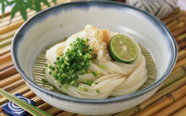 HD PC desktop wallpaper of a bowl of cold udon pasta topped with chopped green onions, grated radish, and a lime slice, set on a bamboo mat with chopsticks.