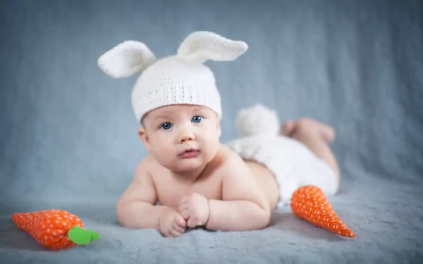 A charming baby in a white bunny hat and diaper lies on a soft blue backdrop, surrounded by orange carrot toys, captured in high-quality photography for a 4K Ultra HD desktop wallpaper.