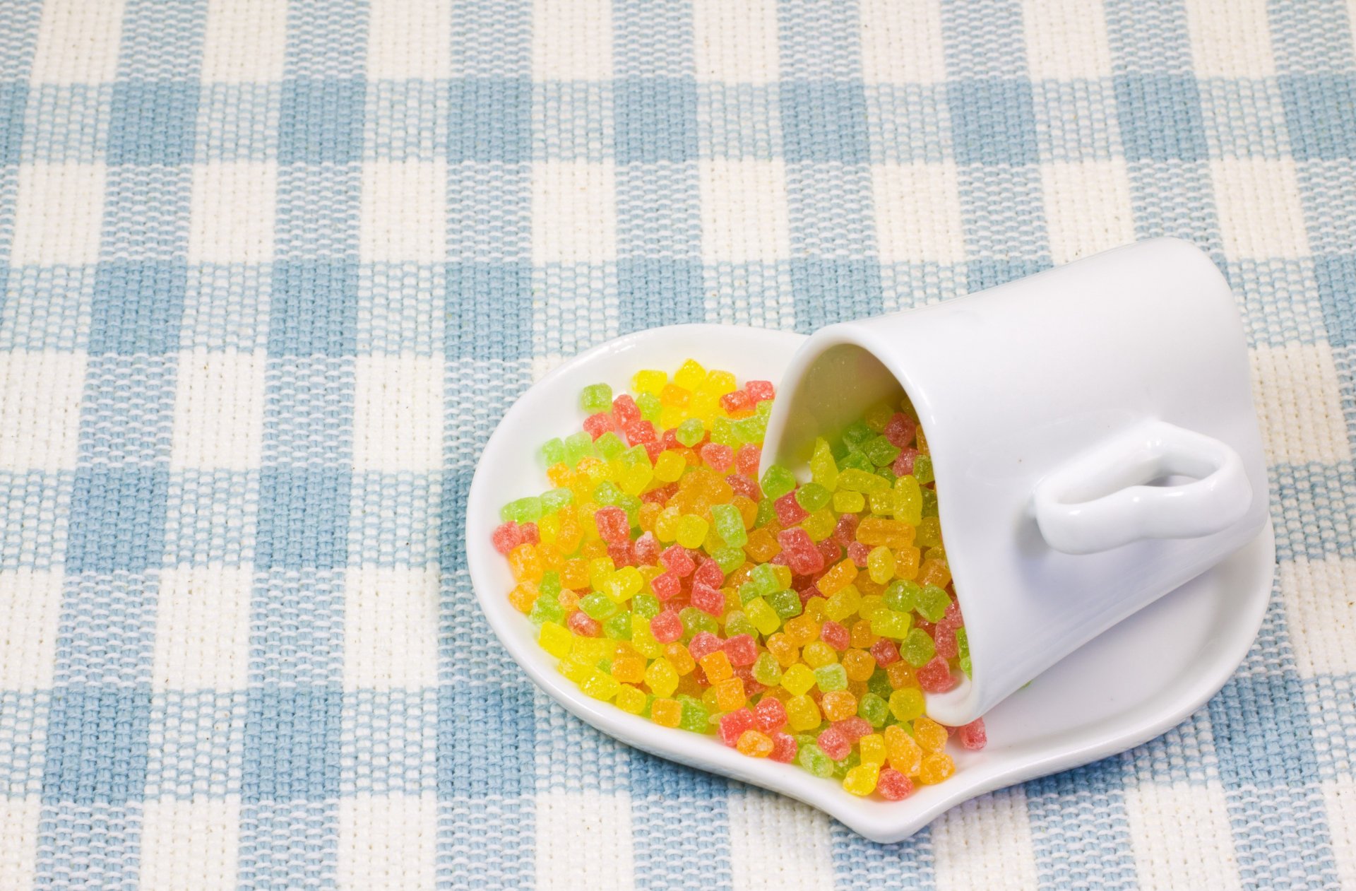 A vibrant display of colorful candy spilling from a white cup onto a heart-shaped dish, set against a blue and white checkered background. A delightful 4K Ultra HD desktop wallpaper.