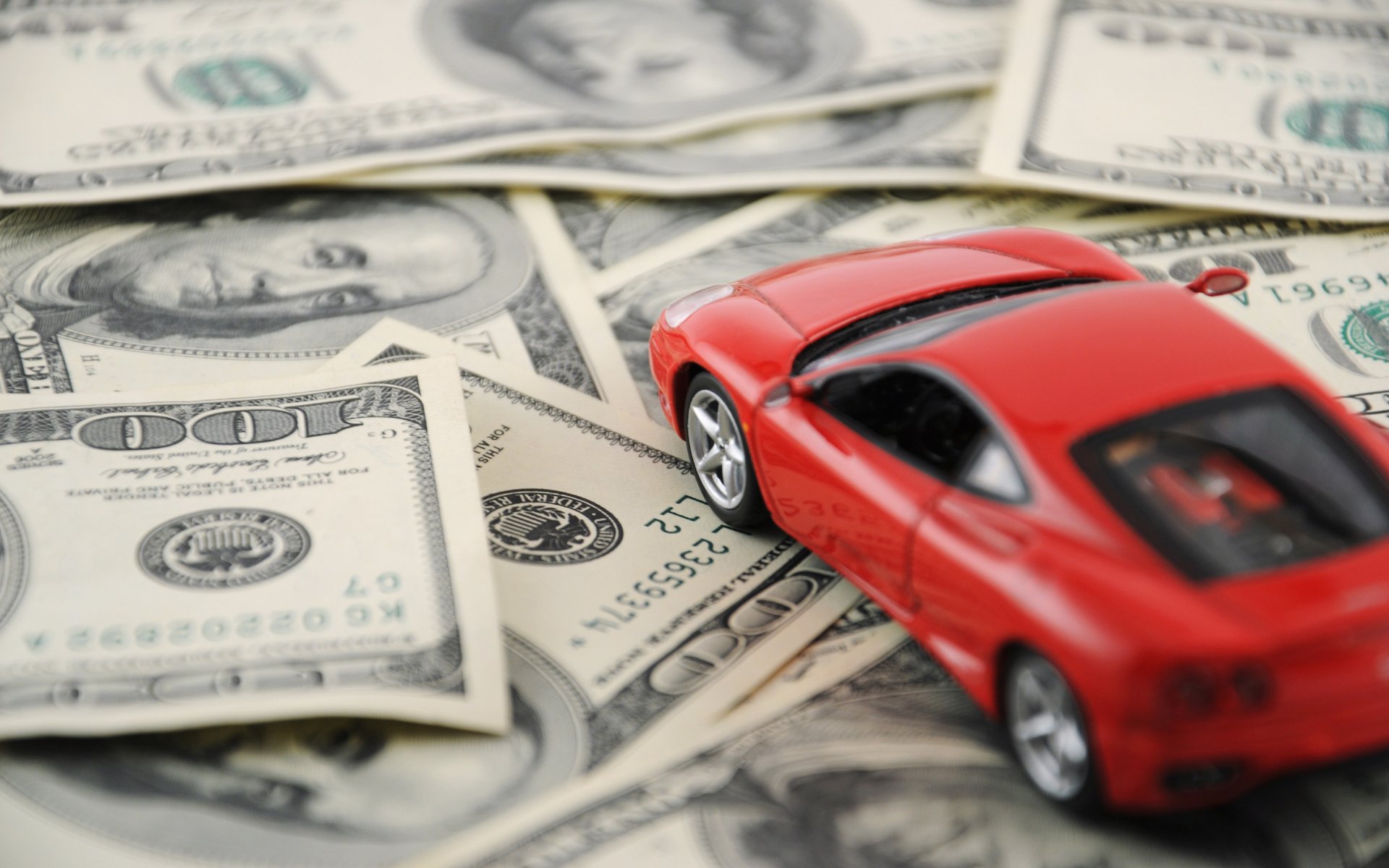 A vibrant red toy car rests on a spread of hundred-dollar bills, showcasing a dynamic contrast between wealth and luxury in this HD desktop wallpaper.