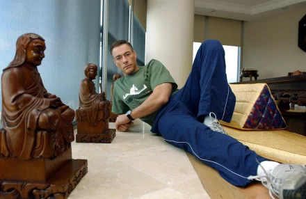 Jean-Claude Van Damme relaxes indoors, dressed casually, with wooden sculptures around him, showcasing a blend of martial arts and tranquility in this HD wallpaper.