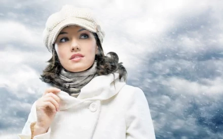 A beautiful woman dressed in a stylish white coat and hat stands against a backdrop of cloudy skies and falling snow, creating a striking HD desktop wallpaper.