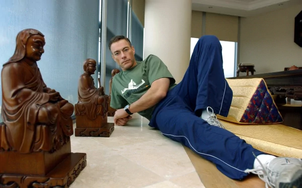 Jean-Claude Van Damme relaxes indoors, dressed casually, with wooden sculptures around him, showcasing a blend of martial arts and tranquility in this HD wallpaper.