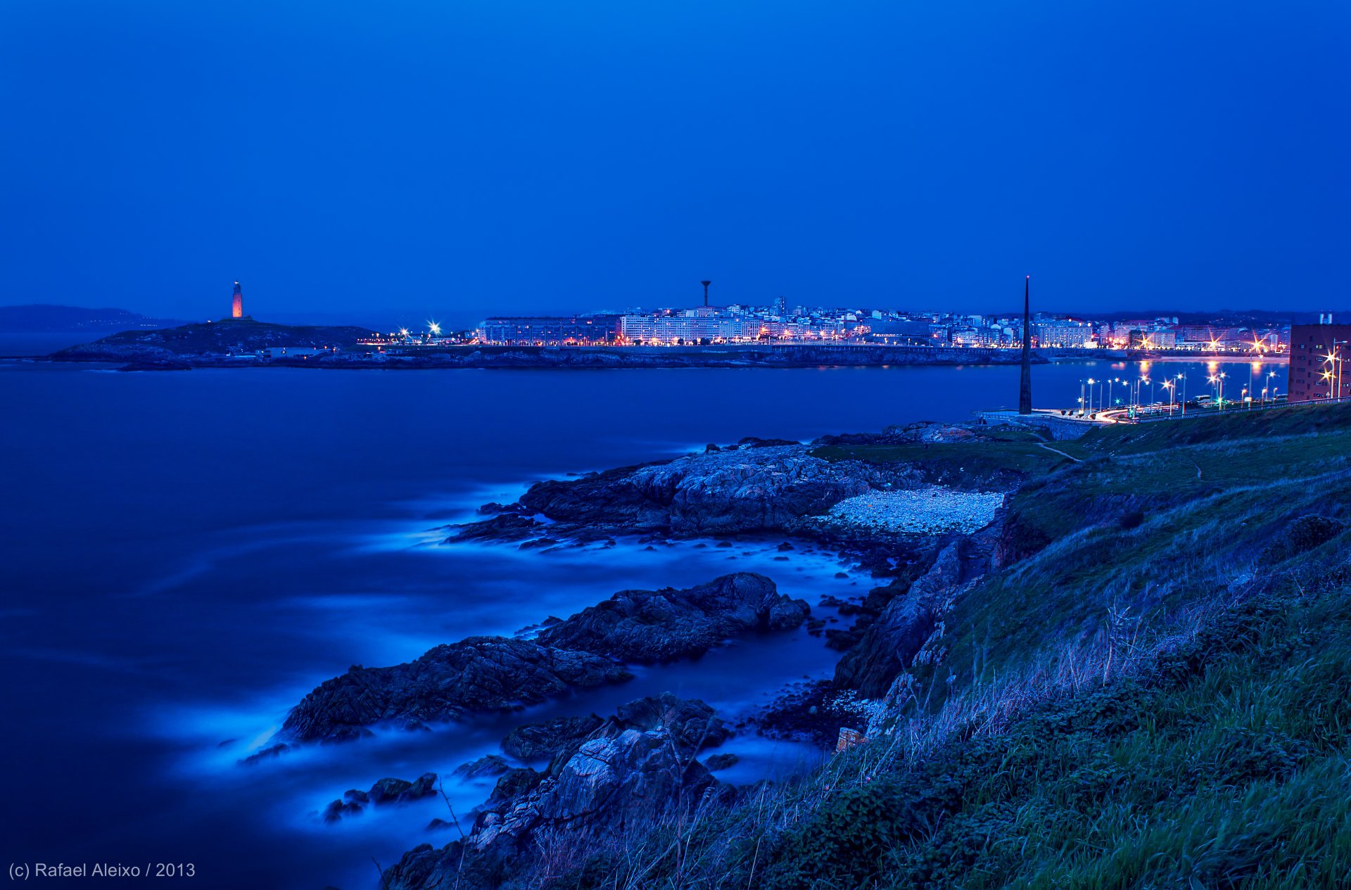 A serene coastal cityscape at dusk, showcasing shimmering lights reflecting on the water, contrasting with the rugged shoreline in a stunning HD wallpaper background.