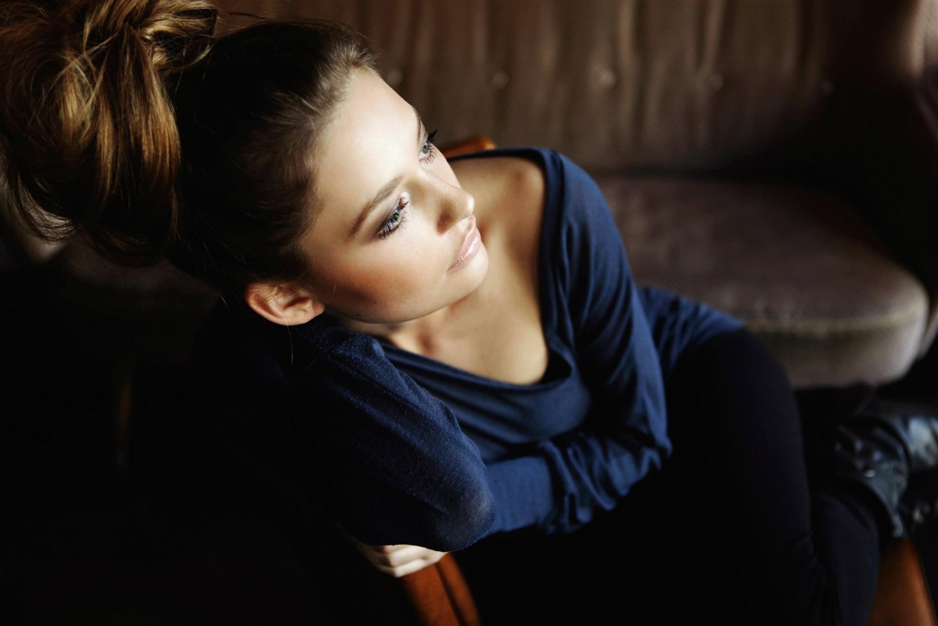HD desktop wallpaper featuring a beautiful woman with styled hair in a dark top, gazing thoughtfully while seated indoors.