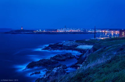 A serene coastal cityscape at dusk, showcasing shimmering lights reflecting on the water, contrasting with the rugged shoreline in a stunning HD wallpaper background.