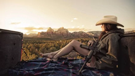 A woman in a cowboy hat relaxes outdoors, seated on a blanket with a gun in hand, set against a stunning sunset and rugged landscape, creating an evocative Girls & Guns scene.