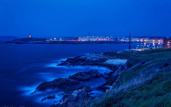 A serene coastal cityscape at dusk, showcasing shimmering lights reflecting on the water, contrasting with the rugged shoreline in a stunning HD wallpaper background.