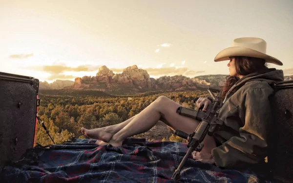 A woman in a cowboy hat relaxes outdoors, seated on a blanket with a gun in hand, set against a stunning sunset and rugged landscape, creating an evocative Girls & Guns scene.