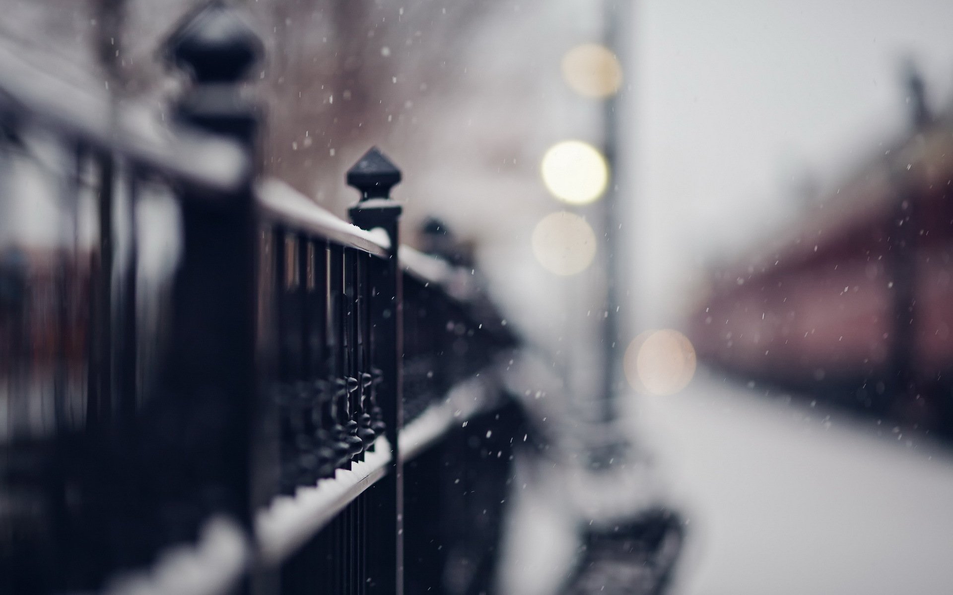 HD desktop wallpaper showing a close-up of a man-made metal fence with a soft focus urban background and falling snow.