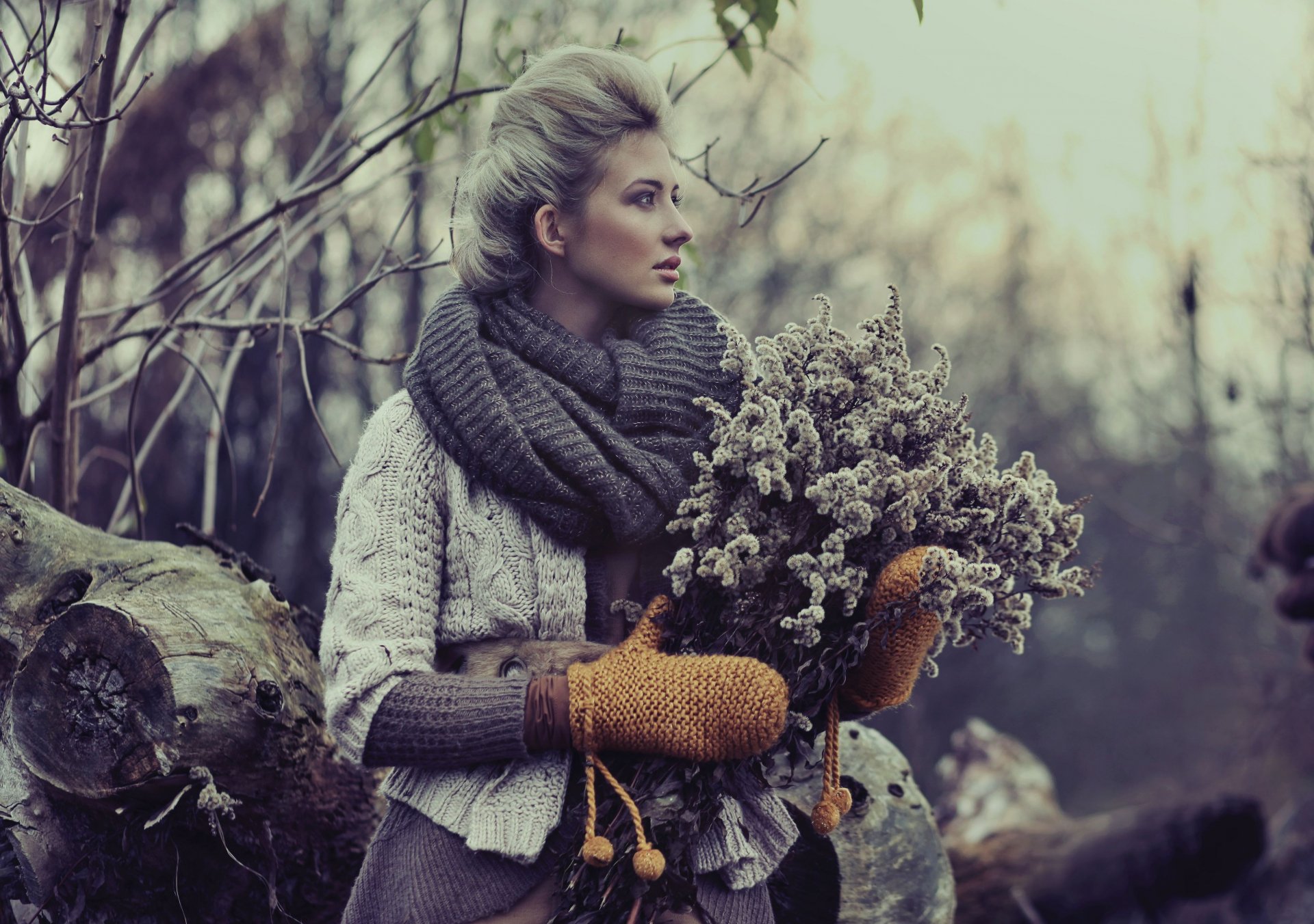 A woman in a cozy outfit, wearing warm mittens and a large scarf, stands in a serene outdoor setting while holding a bundle of dried flowers. A beautiful HD desktop wallpaper capturing a reflective mood.