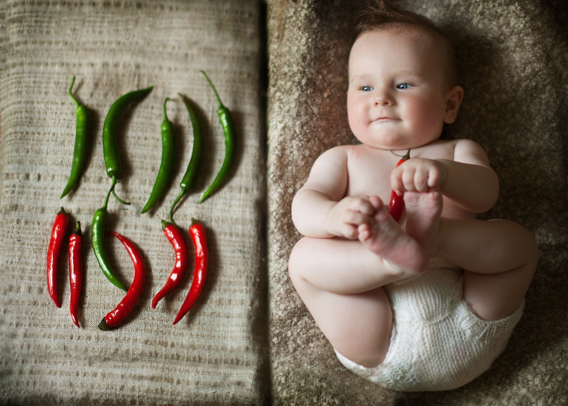 HD photography desktop wallpaper of a smiling baby lying on a textured surface next to an arrangement of red and green chili peppers.