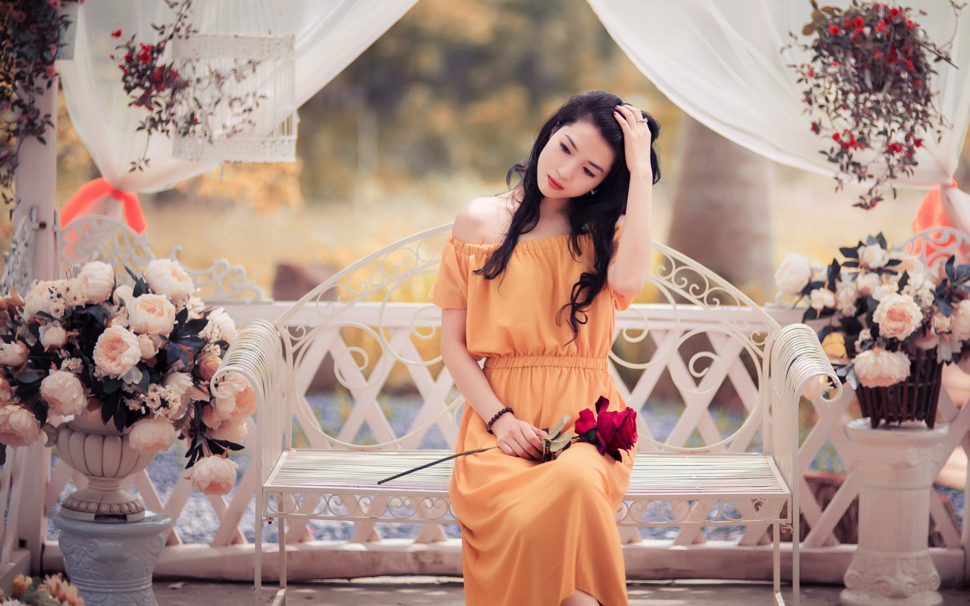 HD desktop wallpaper featuring an Asian woman in an orange dress sitting on a bench, surrounded by flowers and soft white drapery in a tranquil garden setting.