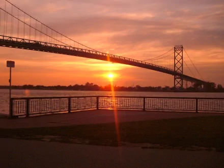 HD PC desktop wallpaper showing the man-made Ambassador Bridge at sunset, silhouetted over the river with sun glinting on the water and a foreground railing.