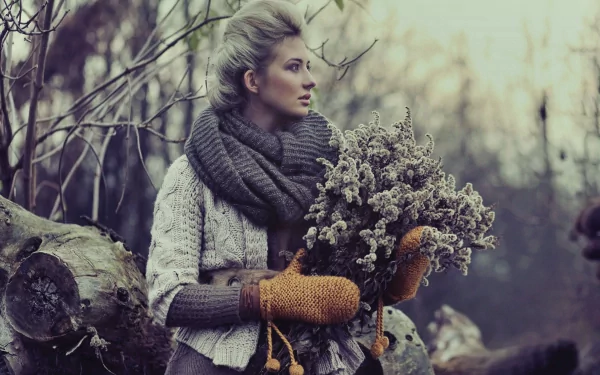 A woman in a cozy outfit, wearing warm mittens and a large scarf, stands in a serene outdoor setting while holding a bundle of dried flowers. A beautiful HD desktop wallpaper capturing a reflective mood.