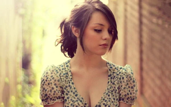 HD desktop wallpaper of a woman, Imogen Dyer, standing outdoors against a blurred background, looking down with soft sunlight filtering through.