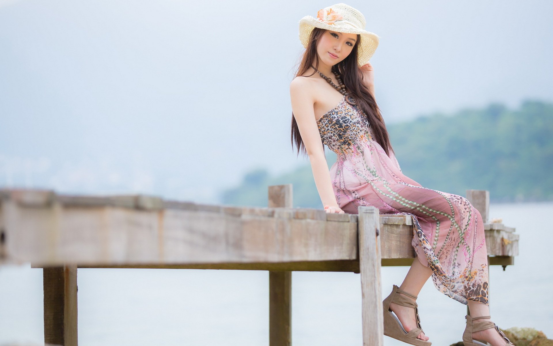 A woman in a light summer dress and wide-brimmed hat sits on a wooden pier by a serene body of water, posing for the camera. This HD desktop wallpaper and background features an Asian woman enjoying a peaceful day outdoors.