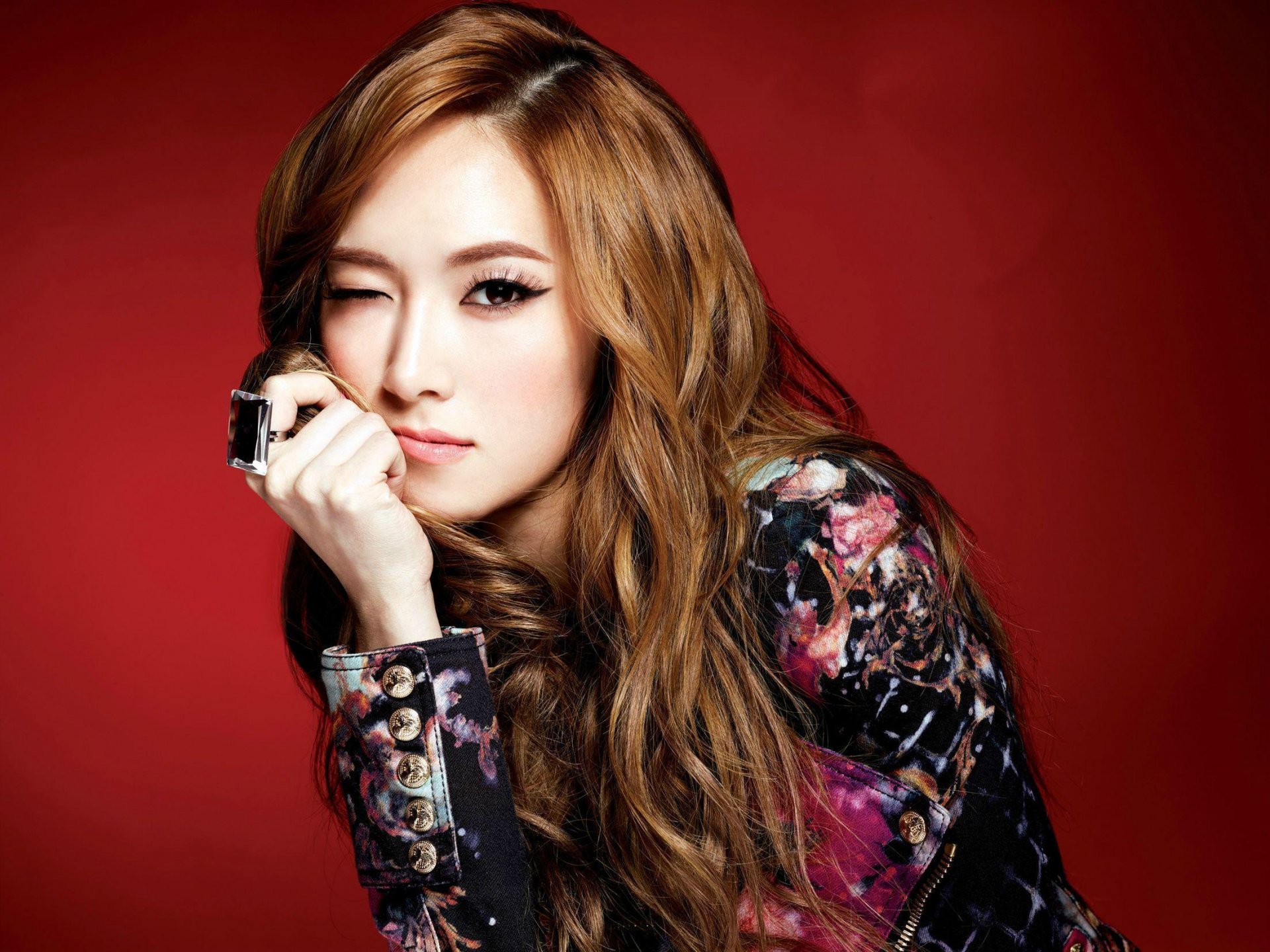 HD desktop wallpaper featuring an Asian woman with long, wavy hair, winking at the camera against a red background. She wears a patterned top and rests her chin on her hand.