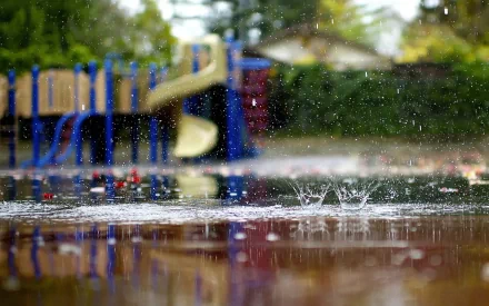 HD desktop wallpaper showing raindrops splashing on a reflective surface with a blurred playground in the background. The image captures the serene beauty of rain.