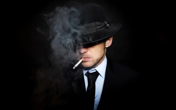 HD desktop wallpaper of a man in a suit and tie, wearing a hat that partially obscures his face. He is smoking a cigarette, with smoke swirling around, creating a moody atmosphere.