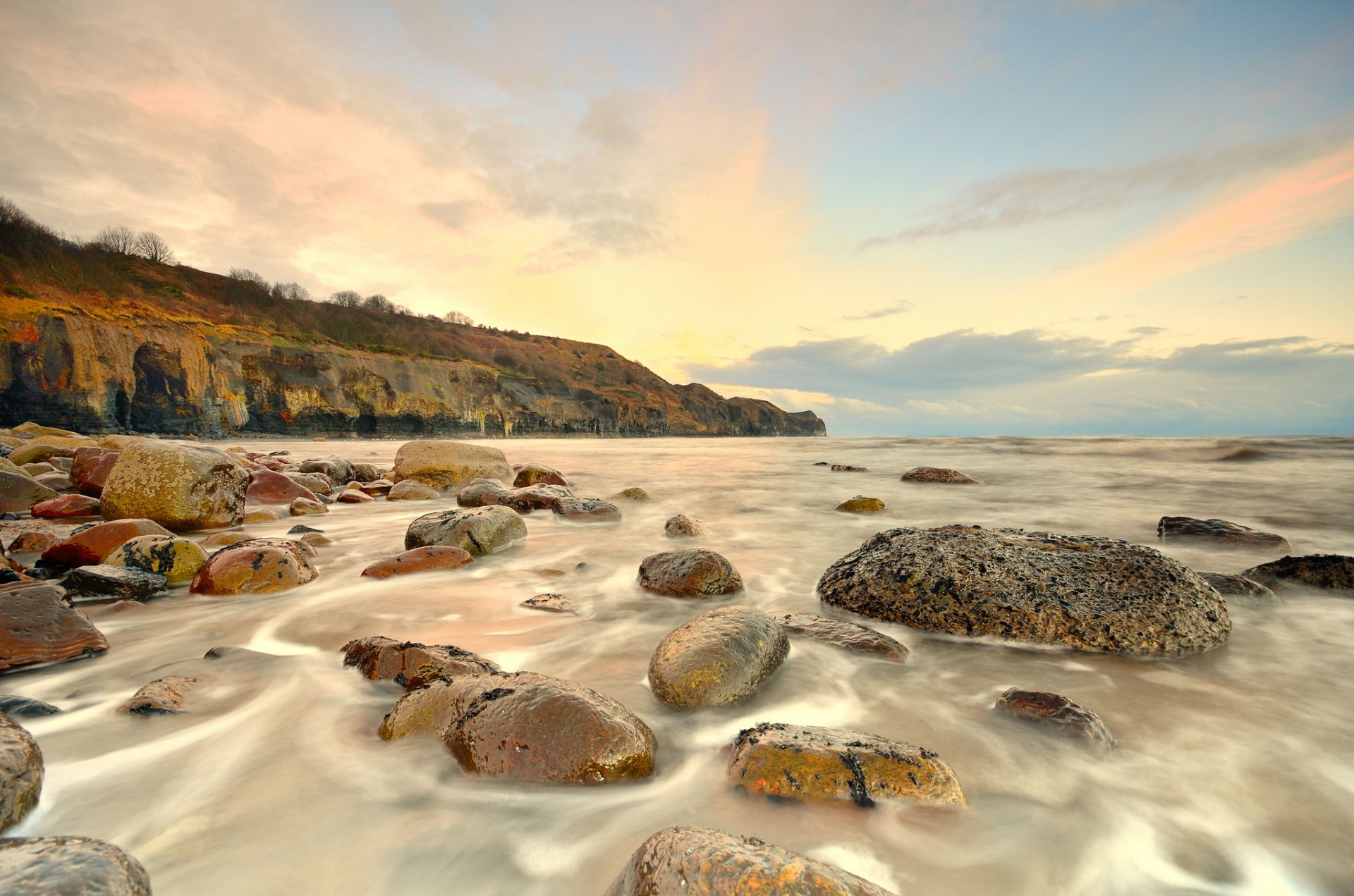 HD PC desktop wallpaper showcasing a serene coastline with smooth rocks, calm waters, and a soft, pastel sky in a natural coastal landscape.