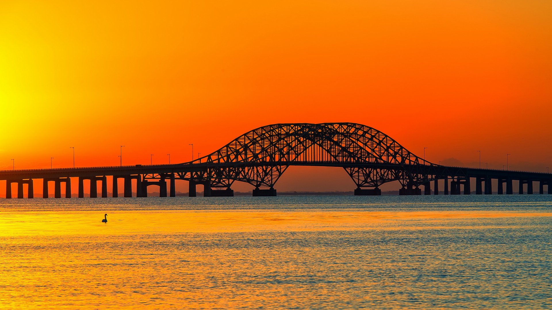 HD PC desktop wallpaper featuring a man-made steel bridge silhouetted against a vibrant orange sunset over calm water.