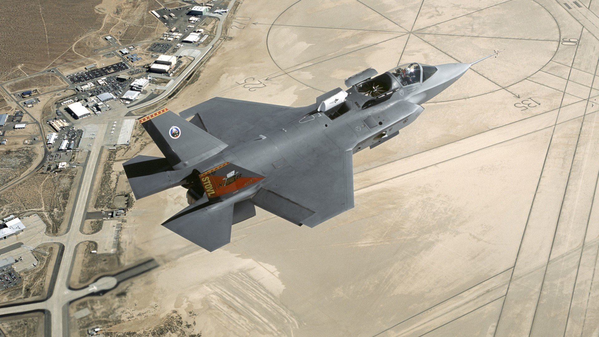 A Lockheed Martin F-35 Lightning II military jet flies over a desert airbase, captured in high definition as a PC desktop wallpaper background.