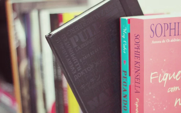 A close-up of colorful books on a shelf, featuring a black book and works by Sophie Kinsella, set against a blurred background. This HD wallpaper adds a literary touch to any desktop.