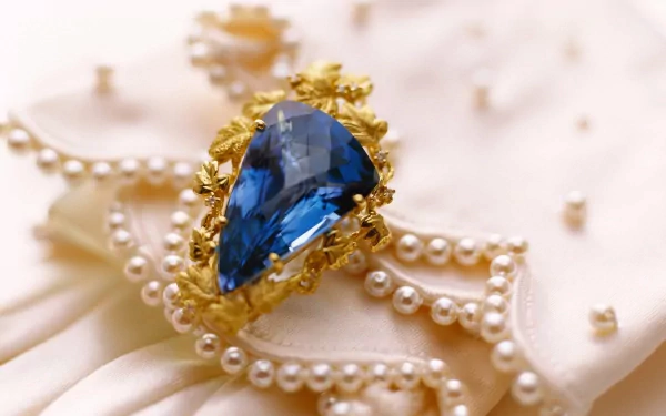 A close-up of elegant jewelry featuring a large blue gemstone set in intricate golden detailing, surrounded by pearl strands on a soft fabric background. HD desktop wallpaper.