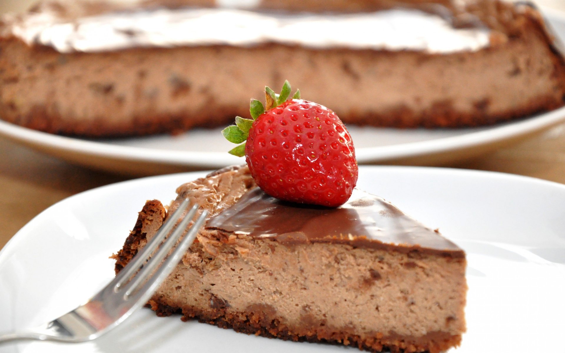HD desktop wallpaper featuring a slice of chocolate cake topped with a fresh strawberry on a white plate, with the whole cake blurred in the background.